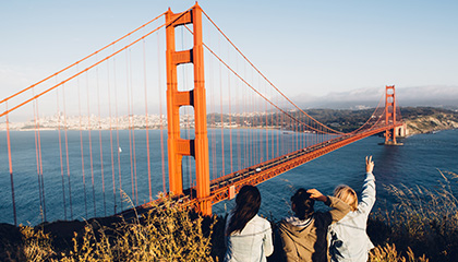 Three individuals stand on a hill, gazing at the Golden Gate Bridge in the distance, framed by a clear blue sky.