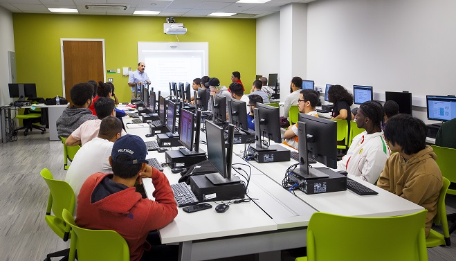 Klasĉambro kun studentoj sidantaj ĉe komputilaj skribotabloj, fronte al instruisto donanta prezenton sur ekrano. La ĉambro havas verdajn murojn kaj modernan dekoracion.