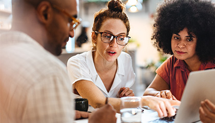 Three individuals collaborating and discussing while looking at a laptop screen together.