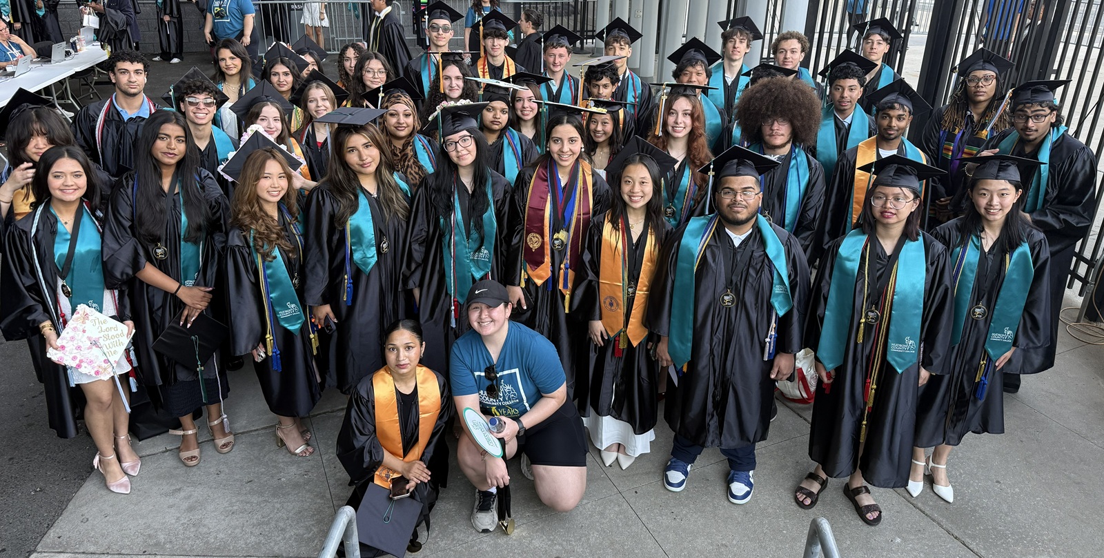 A large group of diverse college graduates in black academic regalia pose for a group photo outdoors at a commencement ceremony. The graduates wear matching teal and black stoles over their gowns, with several individuals also wearing additional honor cords and medals. In the center foreground, two individuals are kneeling: one in a black gown with an orange stole, and another wearing a blue casual t-shirt and shorts. The background shows an outdoor venue with concrete flooring and a dark metal fence. 